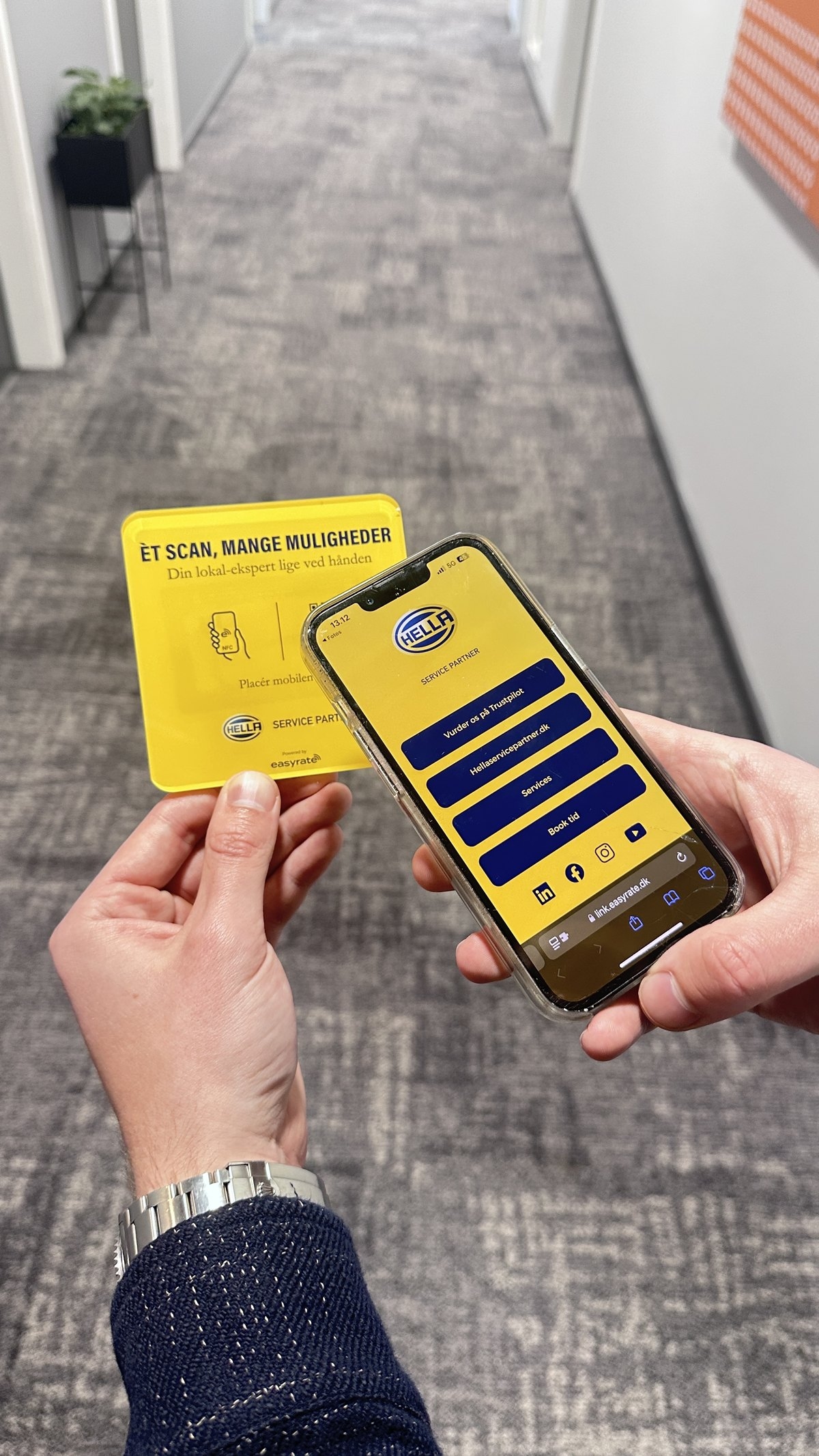 Hand holding a yellow Hella Service Partner NFC plate next to a phone showing the workshop's easylink